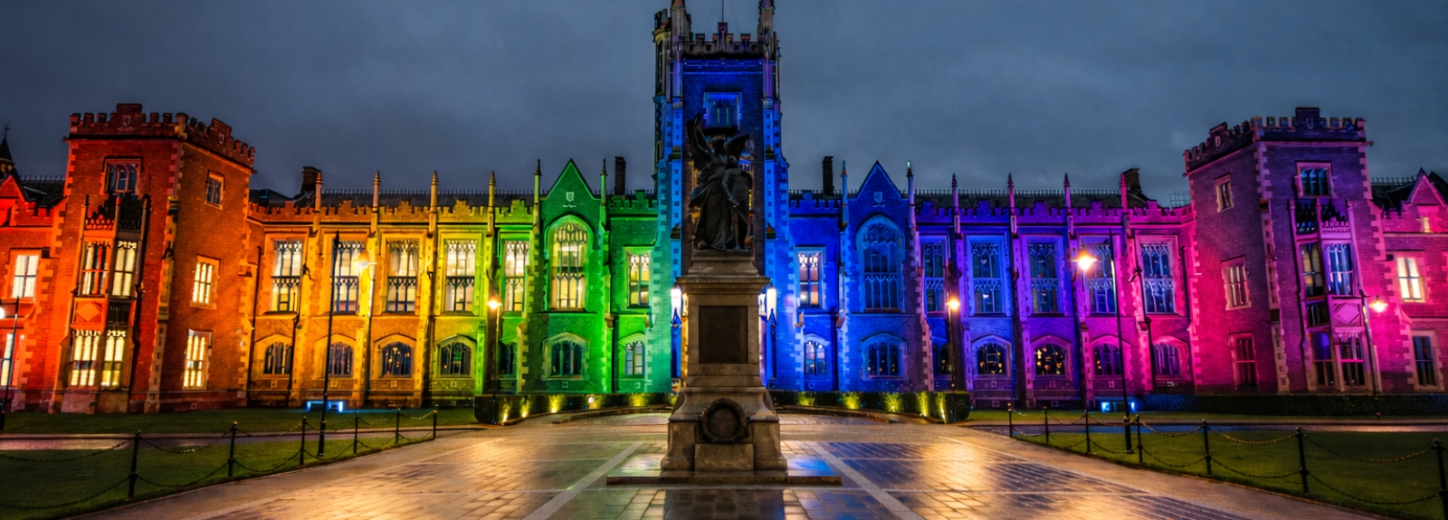Historic university building at night lit in rainbow colours from red and orange to blue, violet and pink, with a central statue and reflections on wet paving